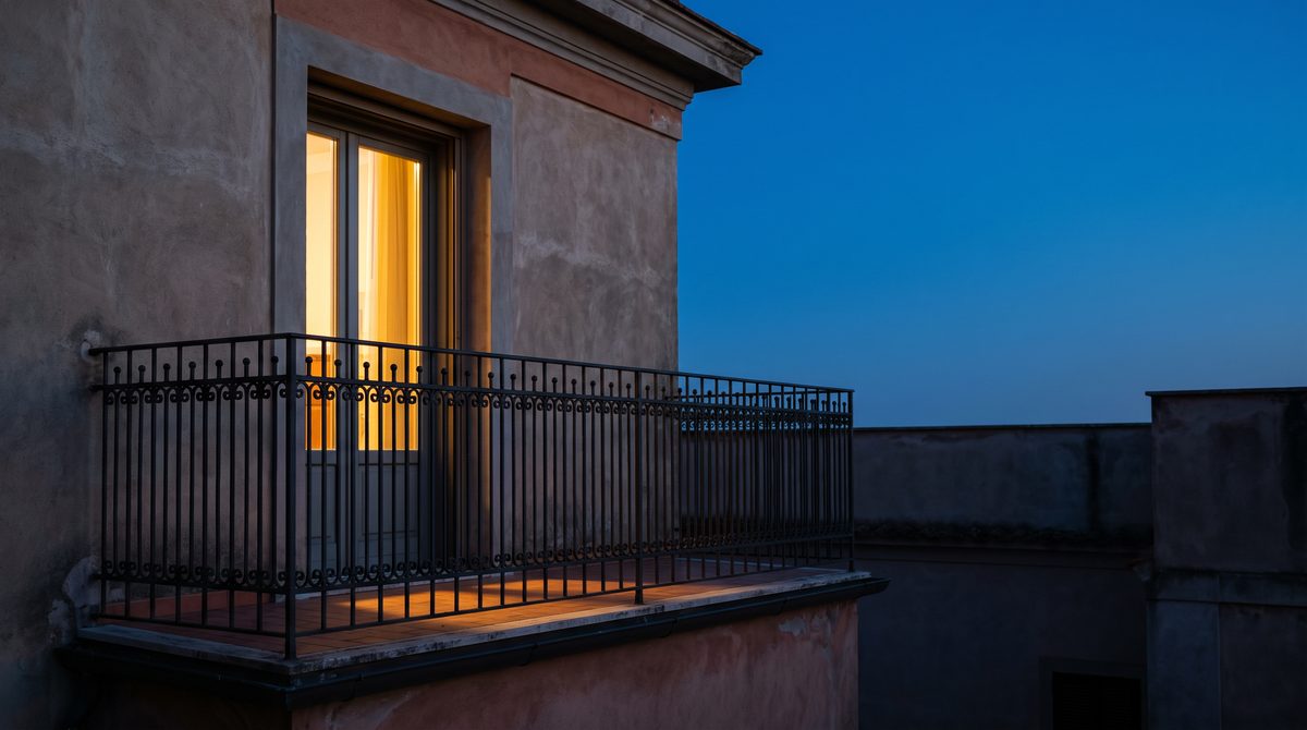 Un balcone con ringhiera in ferro battuto illuminato dalla luce calda di una porta finestra al crepuscolo.