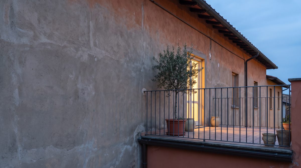 Una facciata di un edificio con un balcone e un albero di ulivo in vaso, illuminato dalla luce di una finestra al crepuscolo.