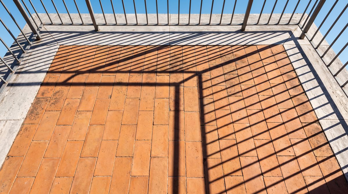 Vista dall'alto di un balcone con pavimento in mattoni rossi e ombre geometriche proiettate dalla ringhiera metallica.