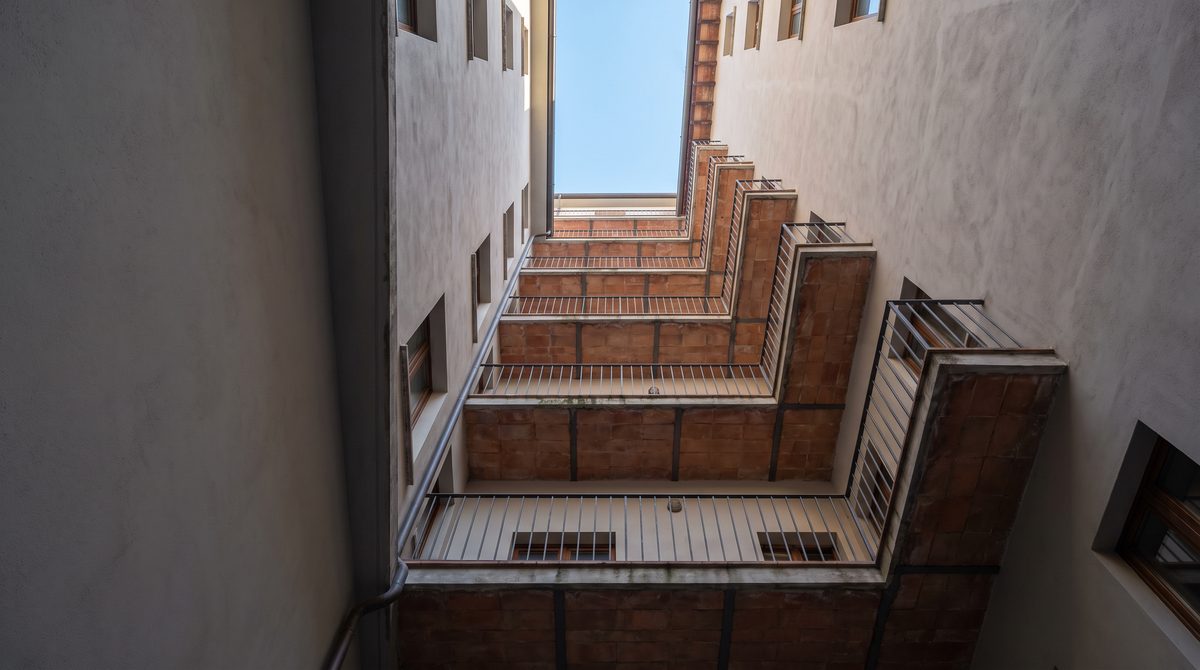 Una vista dal basso verso l'alto di un cortile interno di un edificio, con balconi a zig-zag e il cielo azzurro visibile.