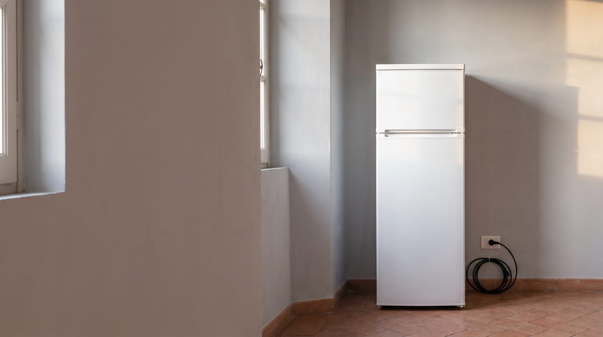 Un frigorifero bianco è posizionato in un angolo di una stanza con pareti grigie, una finestra e un pavimento in cotto.